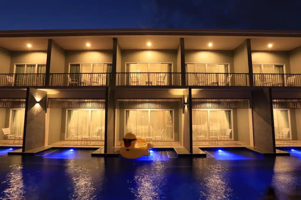 โรงแรมเชียงรายที่ทันสมัยและสว่างไสว พร้อมระเบียงที่มองเห็นสระว่ายน้ำในยามค่ำคืน; เป็ดยางสีเหลืองลอยอยู่บนผิวน้ำในเบื้องหน้า.