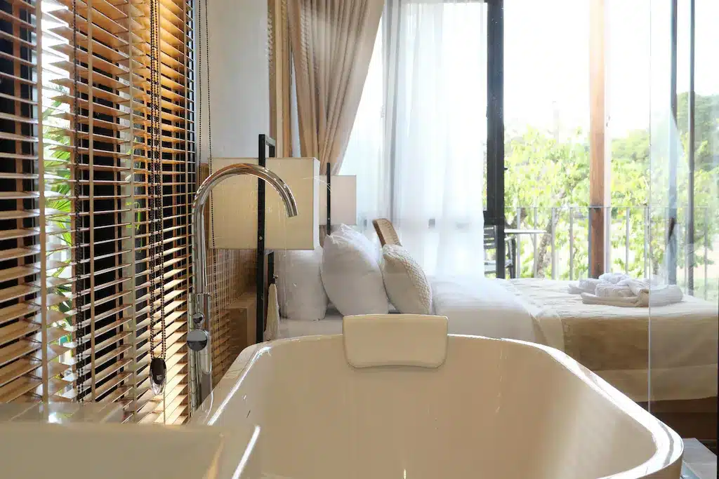 ห้องพักโรงแรมสมัยใหม่ที่รีสอร์ท น่าน ประกอบด้วยอ่างอาบน้ำแบบลอยตัวสีขาวใกล้เตียงที่มีผ้าปูที่นอนสีเบจ หน้าต่างบานใหญ่ และม่านไม้ ช่วยให้แสงธรรมชาติส่องเข้ามาเติมเต็มพื้นที่ได้อย่างเต็มที่.