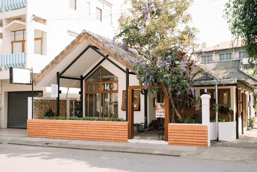 คาเฟ่เล็กๆ ที่มีหน้าต่างบานใหญ่ หลังคากระเบื้อง และต้นไม้อันมีดอกสีม่วงอยู่ด้านหน้า ตั้งอยู่บนถนนในเมืองที่เงียบสงบใกล้ที่พักแสนสบายในเชียงราย.