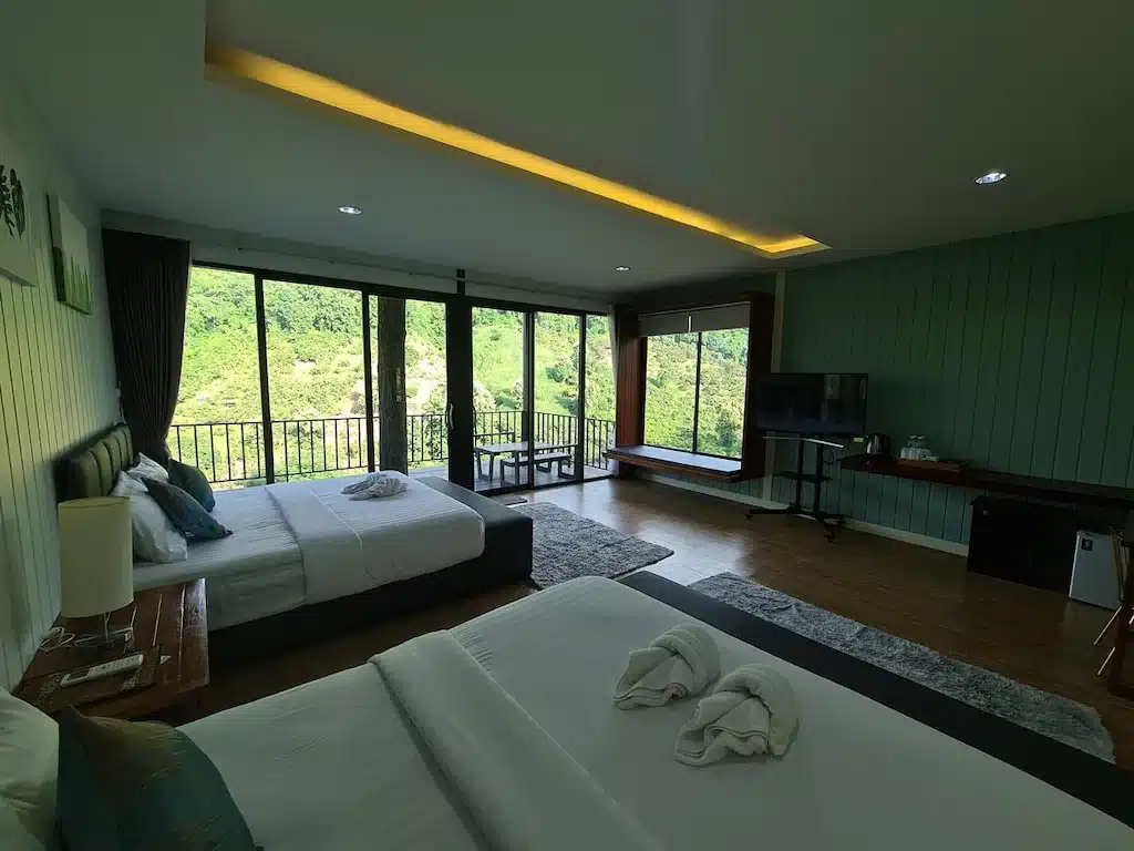 ห้องพักโรงแรมน่านที่กว้างขวางพร้อมเตียงสองเตียง โทรทัศน์ โต๊ะทำงาน หน้าต่างบานใหญ่ และระเบียงที่มองเห็นวิวเนินเขาสีเขียวและต้นไม้.