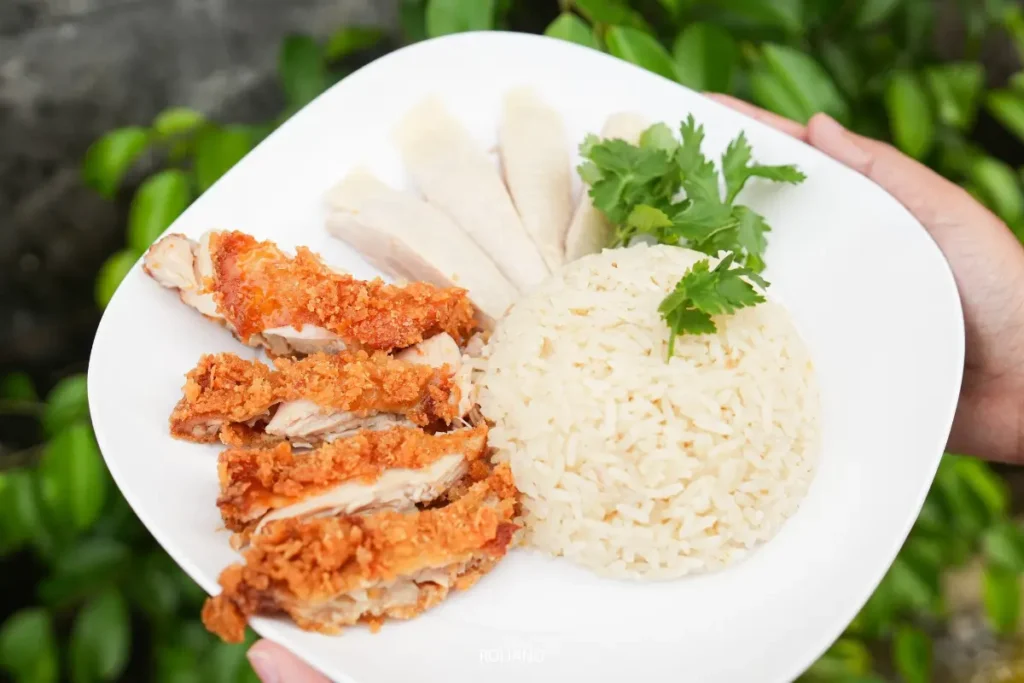 จานที่มีไก่ทอดเป็นชิ้นๆ เนื้อขาวหั่นบางๆ และข้าวที่ตกแต่งด้วยผักชี—ข้าวมันไก่สิงคโปร์ภูเก็ตแท้ๆ—ถือโดยคนๆ หนึ่งอยู่หน้าพืชพรรณเขียวชอุ่ม.