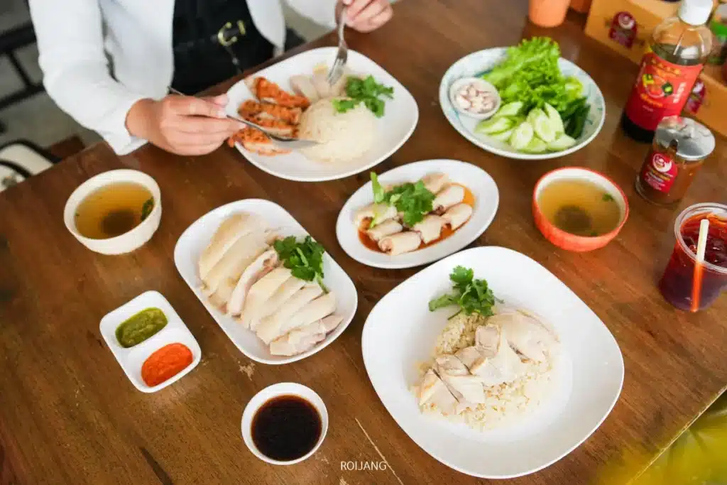 บุคคลหนึ่งกำลังรับประทานอาหารที่โต๊ะไม้ซึ่งจัดวางจานข้าวมันไก่สิงคโปร์ภูเก็ต, แตงกวาหั่น, ซอส, ชามซุป, สลัด และแก้วเครื่องดื่มเย็น.