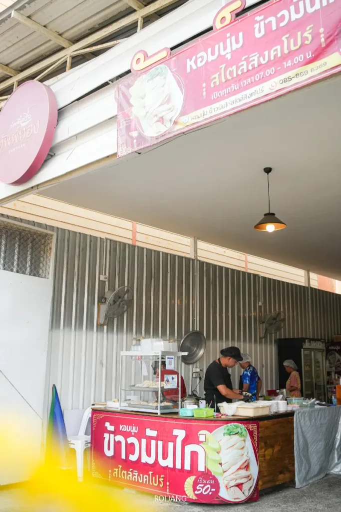 ร้านขายอาหารริมทางที่ขายข้าวมันไก่สิงคโปร์ภูเก็ต โดยมีพ่อค้าแม่ค้าเตรียมอาหารอยู่หลังเคาน์เตอร์ และมีป้ายแสดงรูปภาพของอาหารและราคา ตั้งอยู่ที่ทางเข้าของร้านค้า.