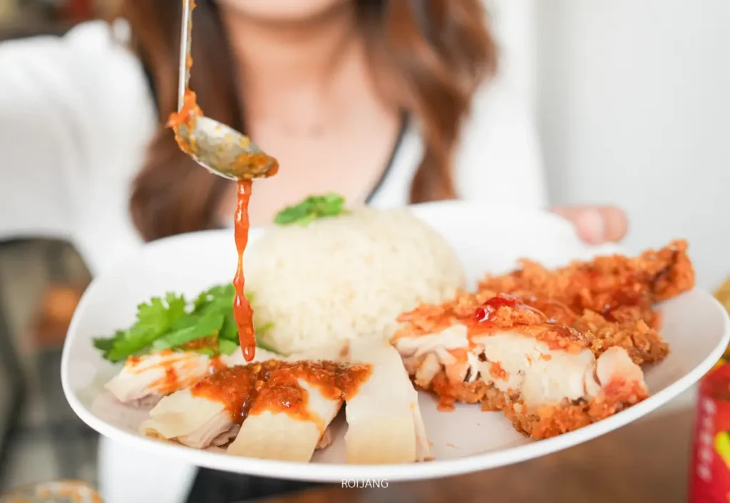 บุคคลหนึ่งถือจานข้าวมันไก่สิงคโปร์ภูเก็ตที่มีข้าวสวย ไก่หั่น ไก่ทอด และผักชีราดซอสจากช้อนลงบนอาหาร.