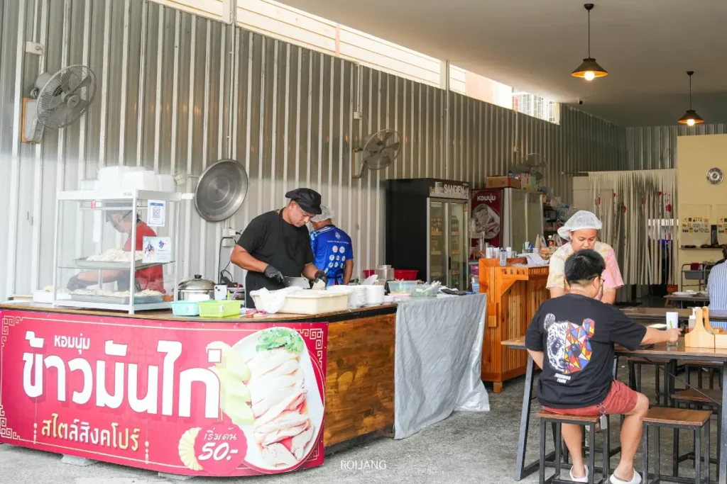 ร้านอาหารไทยขนาดเล็กที่ให้บริการข้าวมันไก่สิงคโปร์ภูเก็ต มีเคาน์เตอร์อาหาร พนักงานสองคนเตรียมอาหาร และลูกค้าที่นั่งอยู่ที่โต๊ะในพื้นที่เปิดโล่งเรียบง่าย.