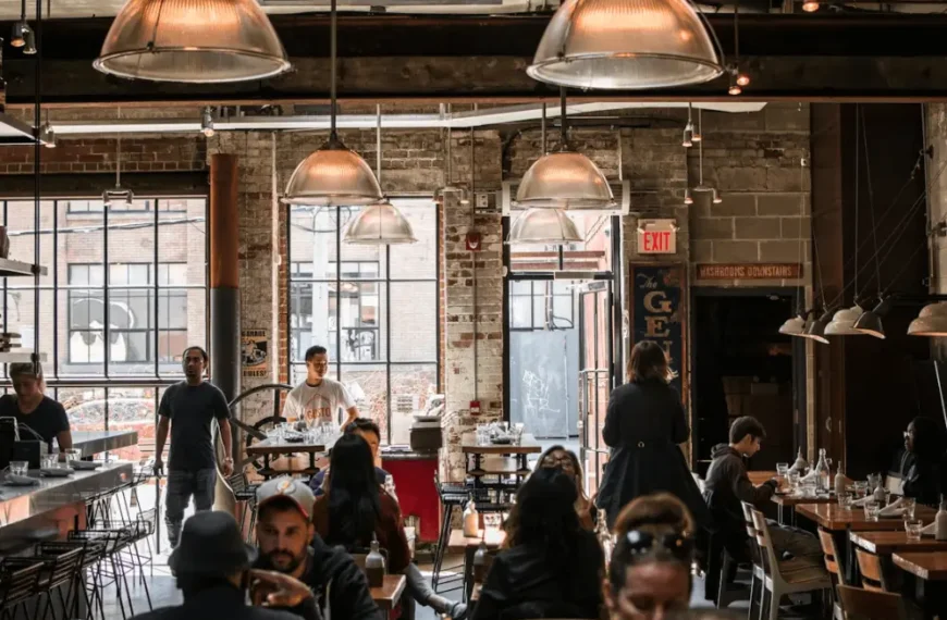 ผู้คนนั่งรับประทานอาหารที่โต๊ะภายในร้านอาหารสไตล์อุตสาหกรรมที่มีผนังอิฐเปลือย หน้าต่างบานใหญ่ และโคมไฟแขวน.