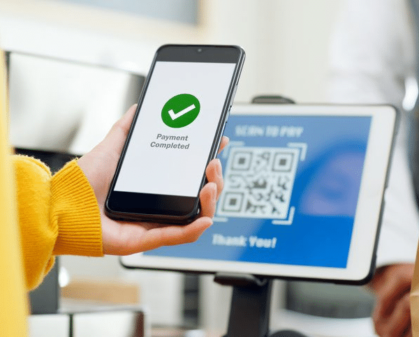 บุคคลหนึ่งถือโทรศัพท์ที่แสดงข้อความ "ชำระเงินเสร็จสมบูรณ์" อยู่ด้านหน้าหน้าจอสแกนคิวอาร์โค้ดสำหรับการชำระเงินที่เคาน์เตอร์ชำระเงิน.