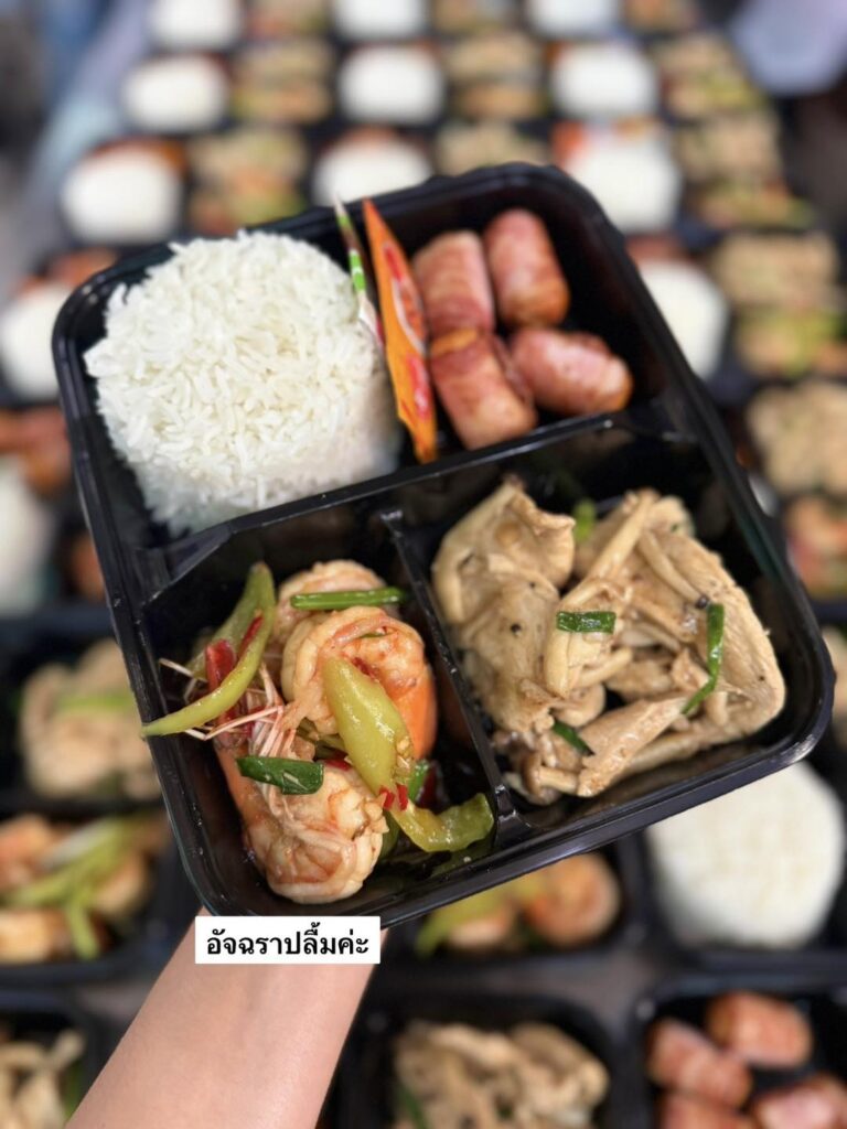 มือหนึ่งถือถาดอาหารสีดำที่มีข้าว ไส้กรอก กุ้งผัด และไก่ผัด ถาดอาหารที่คล้ายกันหลายถาดเบลออยู่ในพื้นหลัง.