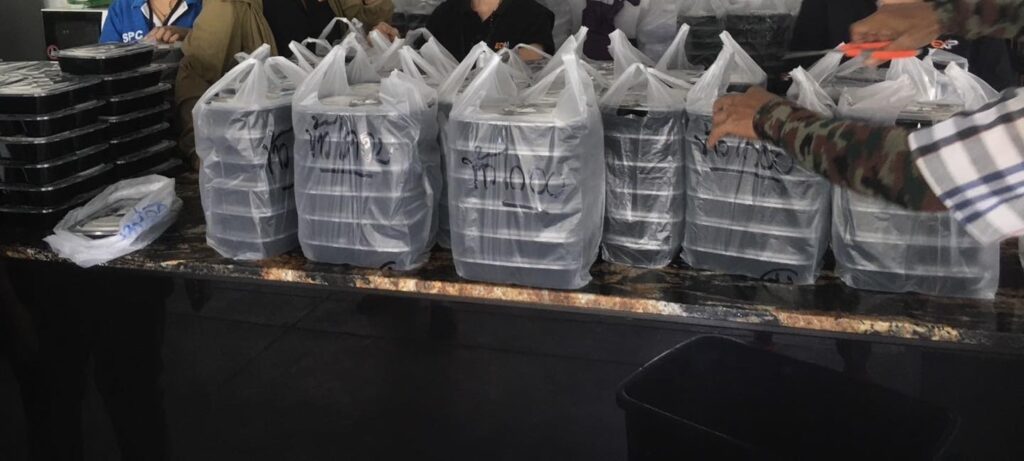 หลายคนกำลังจัดเก็บและจัดเรียงถาดอาหารสีดำซ้อนกันเป็นกองในถุงพลาสติกใสบนเคาน์เตอร์.