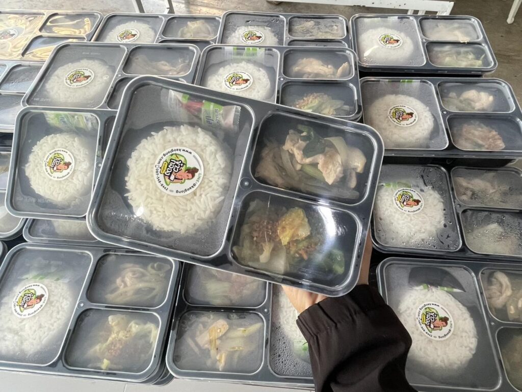 มือหนึ่งถือถาดอาหารบรรจุพร้อมข้าวและกับข้าว; ถาดที่คล้ายกันหลายถาดถูกซ้อนกันอยู่ด้านหลัง.