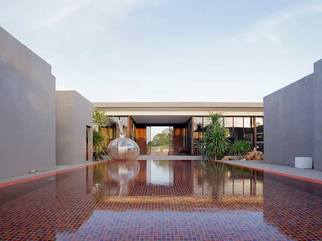 บ้านสมัยใหม่ที่มีหน้าต่างกระจกบานใหญ่และสถาปัตยกรรมแบบมินิมอลสะท้อนอยู่ในสระว่ายน้ำรูปสี่เหลี่ยมผืนผ้าที่ปูด้วยกระเบื้อง ล้อมรอบด้วยภูมิทัศน์ทะเลทราย—โอเอซิสที่ชวนให้นึกถึง ที่เที่ยวเขาใหญ่ สำหรับผู้ที่แสวงหาการพักผ่อนอันเงียบสงบ.