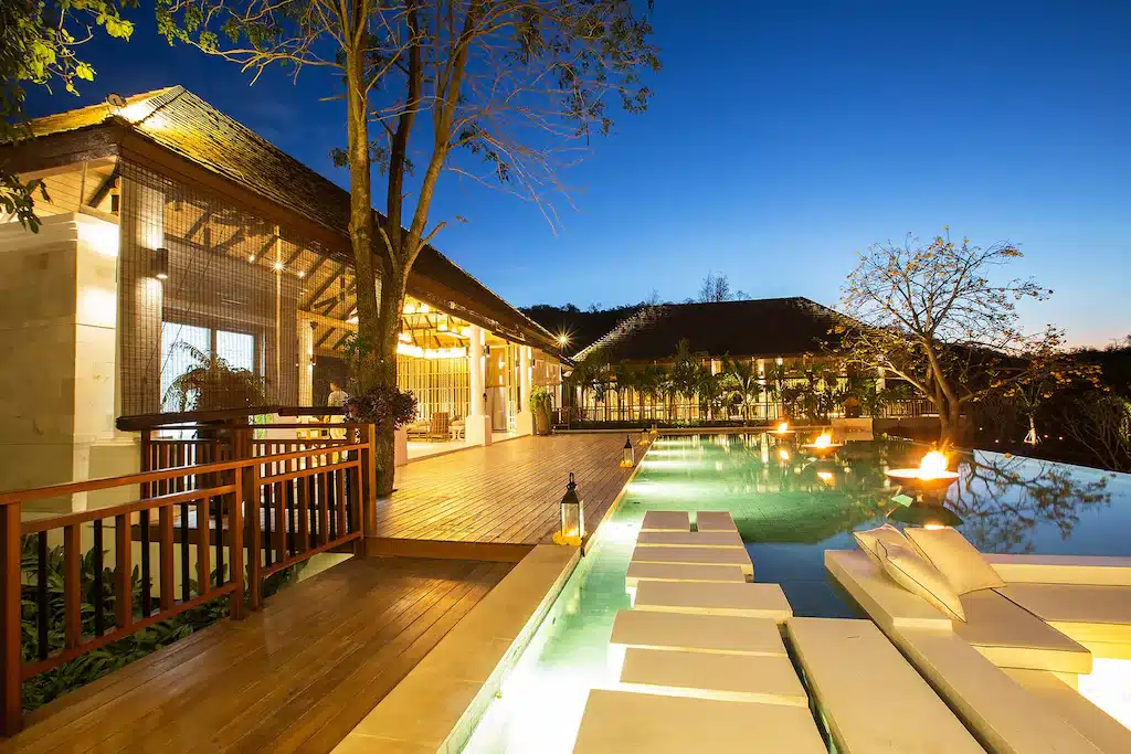 วิลล่าสมัยใหม่พร้อมระเบียงไม้ เก้าอี้เลานจ์ริมสระว่ายน้ำ สระว่ายน้ำแบบอินฟินิตี้ และแสงไฟบรรยากาศยามพระอาทิตย์ตก ท่ามกลางต้นไม้และภูมิทัศน์ที่เขียวชอุ่ม—เหมาะสำหรับผู้ที่กำลังมองหาสถานที่ท่องเที่ยวหรูหราในบรรยากาศเงียบสงบที่เขาใหญ่.