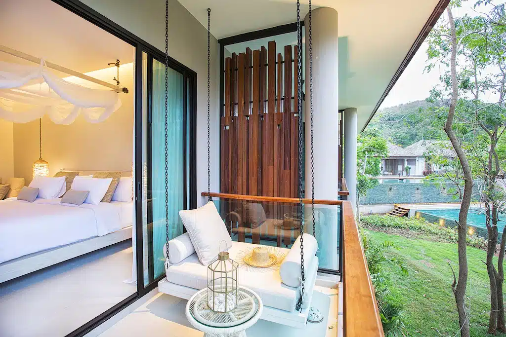 ระเบียงโรงแรมสมัยใหม่พร้อมม้านั่งบุฟองน้ำ หมอน และโต๊ะกลมขนาดเล็ก มองเห็นวิวสีเขียวชอุ่ม เหมาะสำหรับการพักผ่อนหลังจากสำรวจสถานที่ท่องเที่ยวเขาใหญ่ ห้องนอนที่อบอุ่นพร้อมเตียงมุ้งลวดสามารถมองเห็นได้ผ่านประตูกระจก.