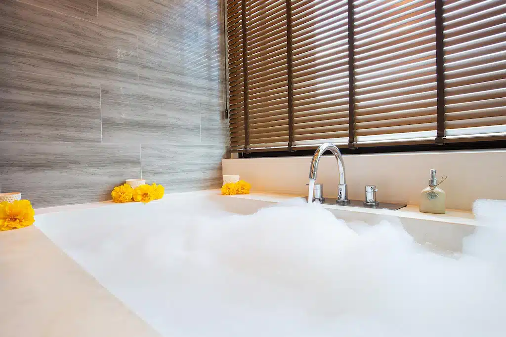 อ่างอาบน้ำที่เต็มไปด้วยฟองสบู่ตั้งอยู่ข้างผนังกระเบื้องสีเทา ล้อมรอบด้วยดอกไม้สีเหลืองและที่จ่ายสบู่—สมบูรณ์แบบหลังจากวันที่ออกไปเที่ยวเขาใหญ่ พร้อมม่านบังตาไม้สีน้ำตาลที่กรองแสงผ่านหน้าต่างอย่างนุ่มนวล.