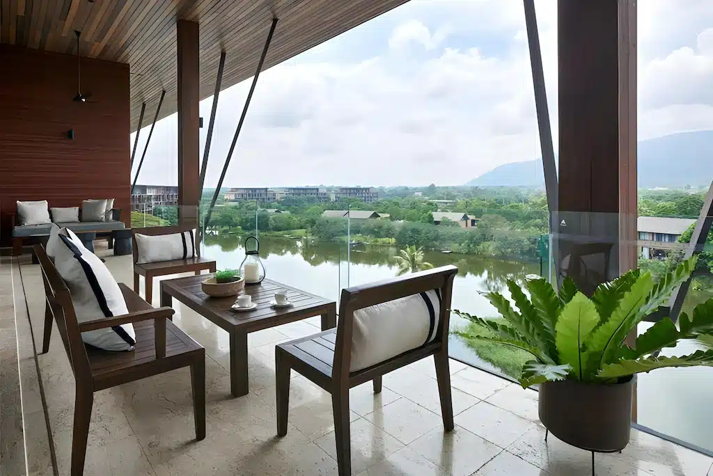 ระเบียงสมัยใหม่พร้อมเก้าอี้ไม้และโต๊ะมองเห็นวิวทะเลสาบและพื้นที่สีเขียว—เหมาะสำหรับการพักผ่อนหลังจากสำรวจสถานที่ท่องเที่ยวเขาใหญ่; ราวระเบียงกระจกและต้นไม้ในกระถางสามารถมองเห็นได้ในเบื้องหน้า.