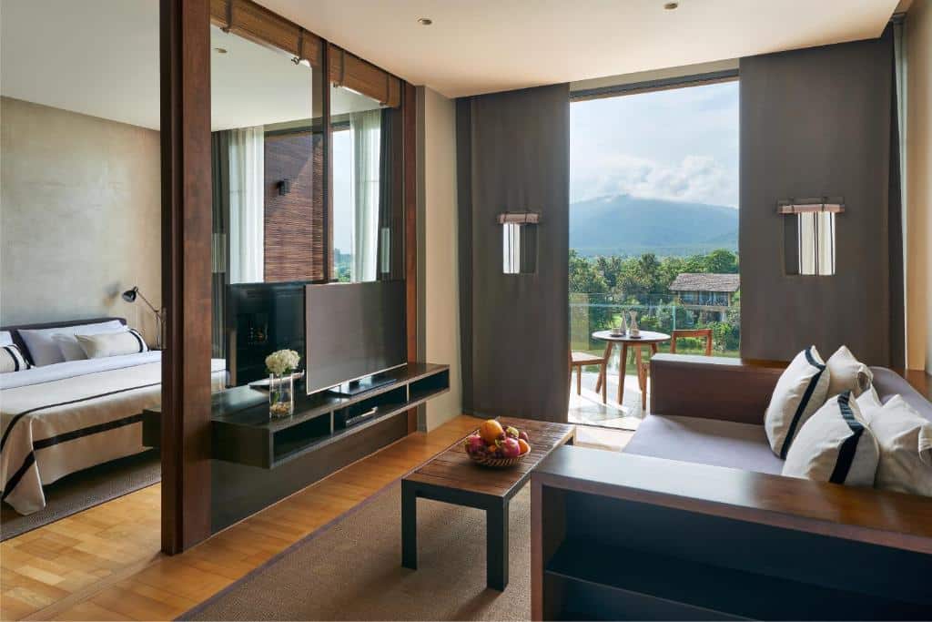 ห้องสวีทโรงแรมสมัยใหม่พร้อมห้องนอนแยก ห้องนั่งเล่น ทีวี ชามผลไม้ และระเบียงที่มองเห็นวิวสีเขียวและภูเขาไกล เหมาะสำหรับการพักผ่อนหลังเที่ยวเขาใหญ่หรือสำรวจสถานที่ท่องเที่ยวยอดนิยมของเขาใหญ่.