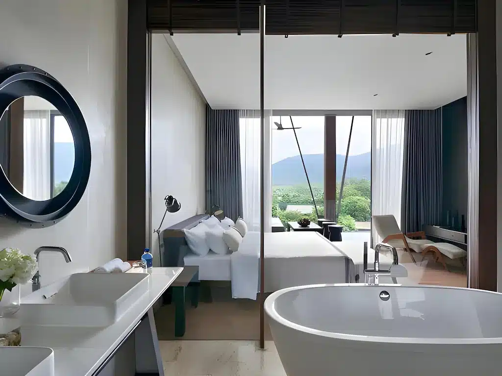 ห้องพักโรงแรมสมัยใหม่พร้อมอ่างอาบน้ำแบบลอยตัว อ่างล้างหน้าคู่ และวิวเตียง โต๊ะทำงาน และหน้าต่างบานใหญ่ที่มองเห็นภูเขาเขียวขจี—เหมาะสำหรับการพักผ่อนหลังจากสำรวจแหล่งท่องเที่ยวเขาใหญ่หรือสถานที่ท่องเที่ยวเขาใหญ่ใกล้เคียง.