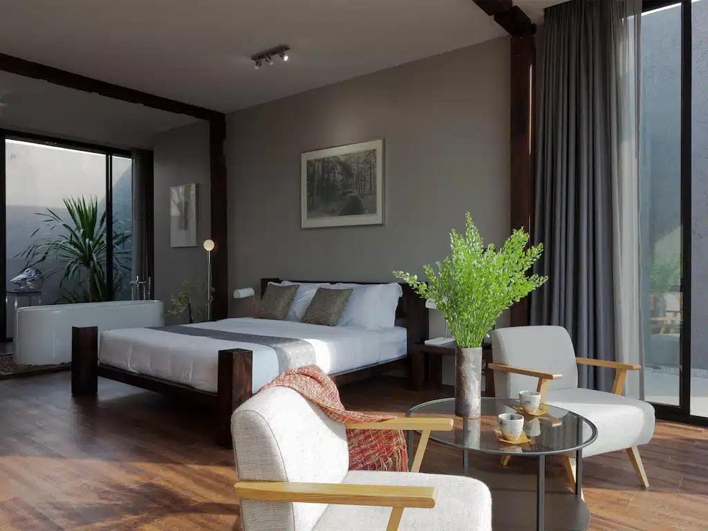 ห้องนอนสมัยใหม่พร้อมพื้นไม้ เตียงขนาดใหญ่ เก้าอี้นวมสองตัวล้อมรอบโต๊ะกาแฟกระจก ต้นไม้ในกระถาง และอ่างอาบน้ำใกล้หน้าต่างที่มีผ้าม่านบาง—เหมาะสำหรับการผ่อนคลายหลังจากสำรวจสถานที่ท่องเที่ยวเขาใหญ่.