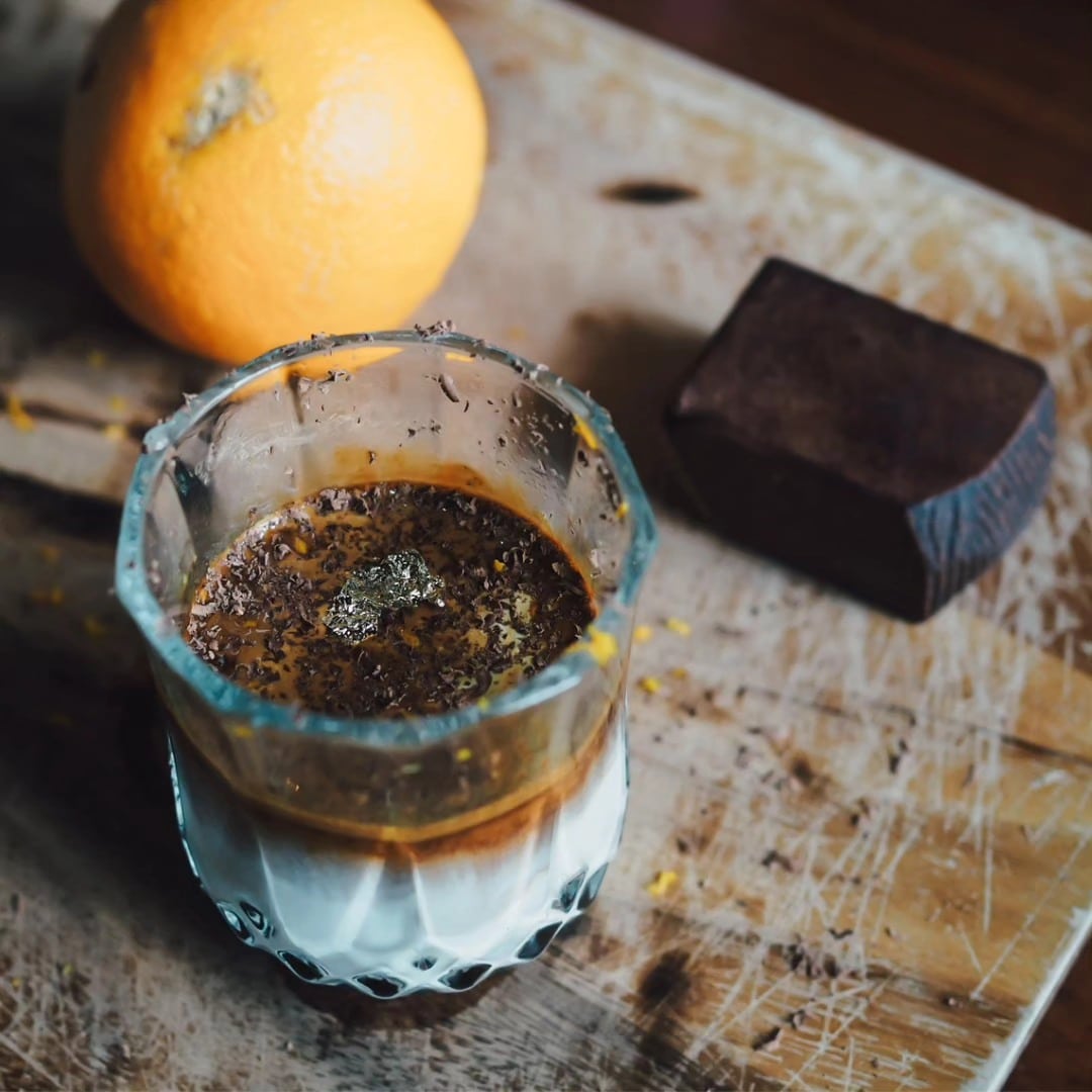 แก้วที่มีกาแฟและนมเป็นชั้นๆ โรยหน้าด้วยช็อกโกแลตขูด วางอยู่บนกระดานไม้ข้างๆ ส้มและช็อกโกแลตหนึ่งก้อน—เหมาะสำหรับการเติมพลังก่อนออกไปสำรวจสถานที่ท่องเที่ยวเขาใหญ่.