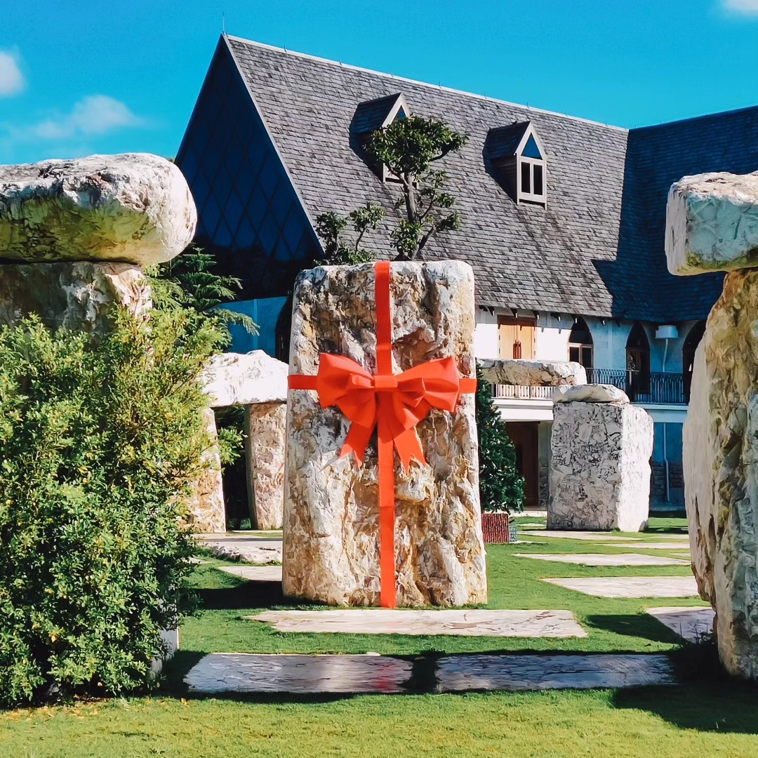 หินสี่เหลี่ยมผืนผ้าขนาดใหญ่ตกแต่งด้วยริบบิ้นสีแดงและโบว์ตั้งอยู่ท่ามกลางแผ่นหินอื่น ๆ บนพื้นที่หญ้า ณ หนึ่งในสถานที่ท่องเที่ยวเขาใหญ่ โดยมีอาคารตั้งอยู่เบื้องหลัง.