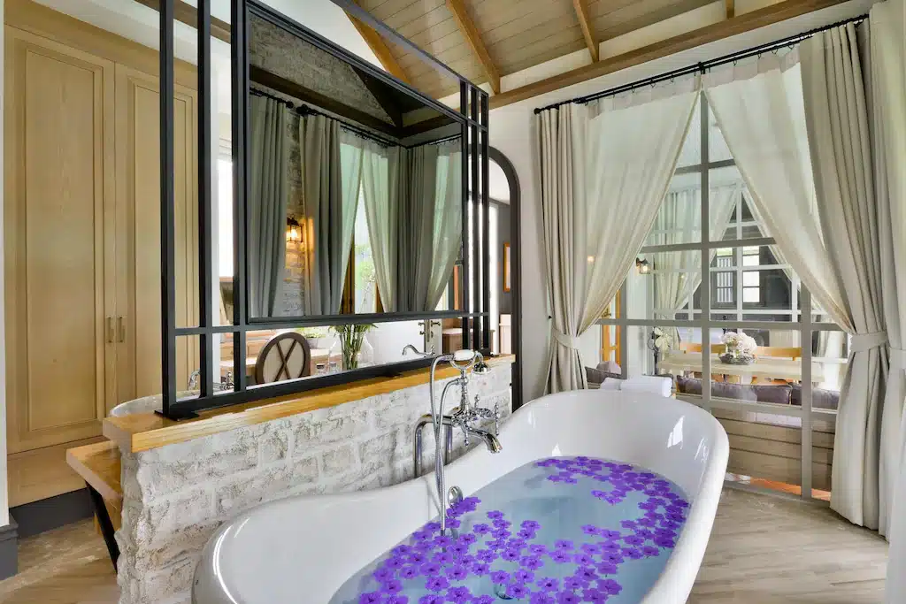 อ่างอาบน้ำสีขาวที่เต็มไปด้วยน้ำและกลีบดอกไม้สีม่วงลอยอยู่ภายในห้องน้ำที่มีสไตล์ พร้อมกระจกบานใหญ่ ผ้าม่าน และของตกแต่งไม้—เหมาะสำหรับการผ่อนคลายหลังจากวันที่ออกไปเที่ยวเขาใหญ่.