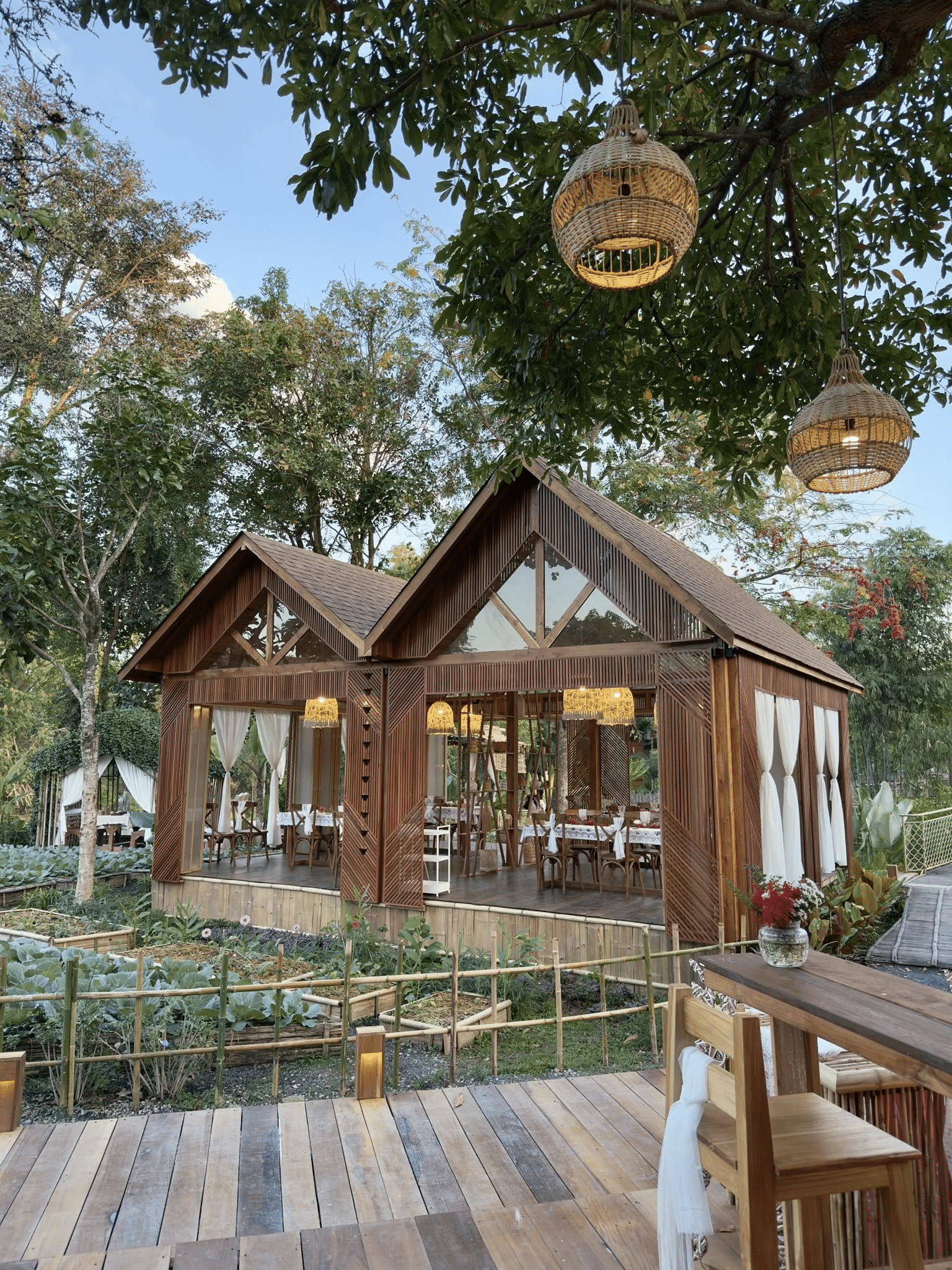 ศาลาไม้เปิดโล่งพร้อมโต๊ะและเก้าอี้ตั้งอยู่ในสวนที่เขียวชอุ่ม ล้อมรอบด้วยต้นไม้และโคมไฟแขวนใต้ท้องฟ้าแจ่มใส—เป็นจุดที่น่าดึงดูดสำหรับการพักผ่อนที่ ที่เที่ยวเขาใหญ่.