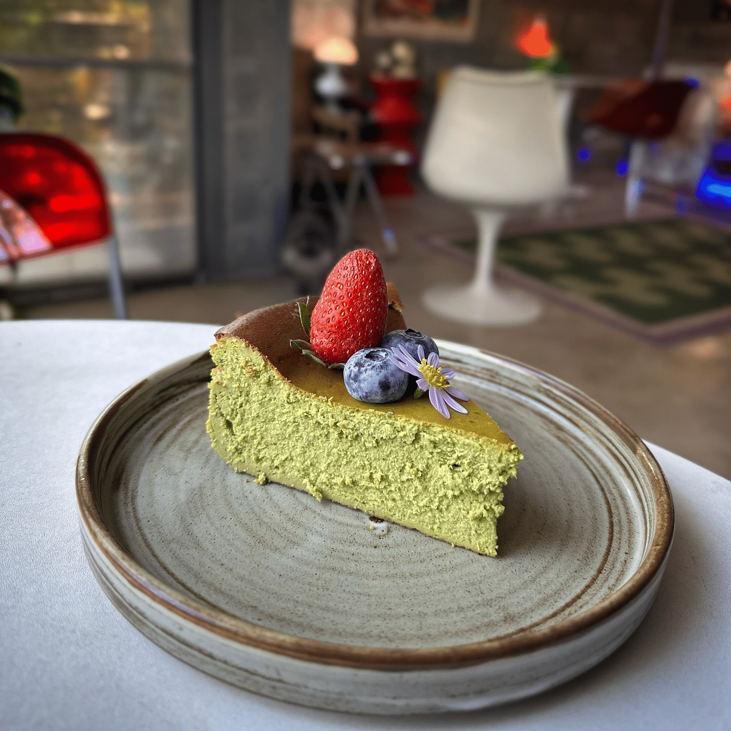 ชีสเค้กสีเขียวชิ้นหนึ่งตกแต่งด้วยสตรอว์เบอร์รี บลูเบอร์รี และดอกไม้ที่รับประทานได้ เสิร์ฟบนจานเซรามิกทรงกลมในบรรยากาศภายในที่ทันสมัย—เหมาะสำหรับการผ่อนคลายหลังจากสำรวจสถานที่ท่องเที่ยวเขาใหญ่.