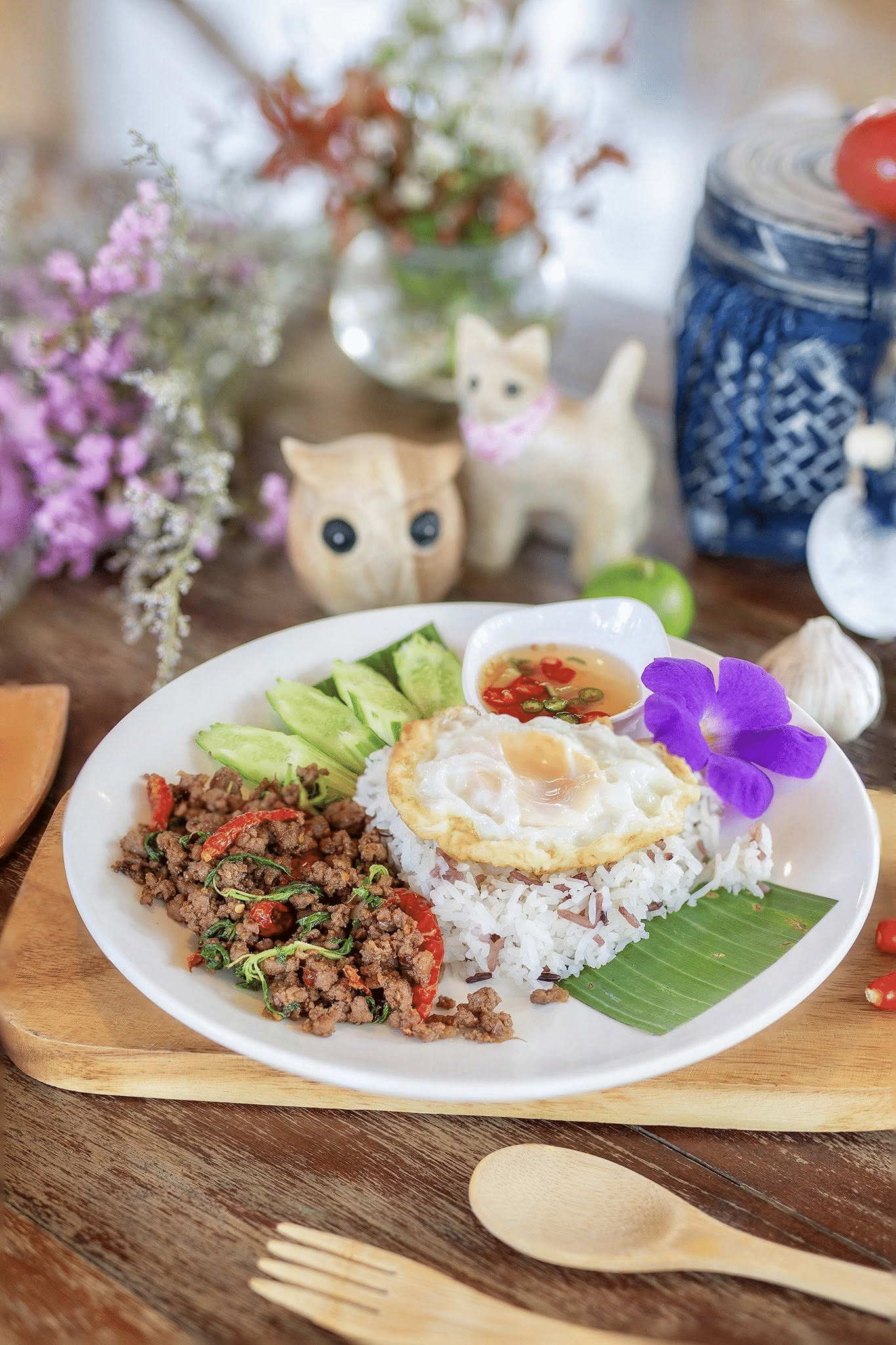 ข้าวสวยหนึ่งจาน ราดด้วยไข่ดาว, เนื้อสับผัดพริกและโหระพา, แตงกวาหั่นแว่น, และน้ำจิ้มถ้วยเล็ก ตกแต่งด้วยดอกไม้สีม่วง—สมบูรณ์แบบหลังจากวันที่สำรวจ ที่เที่ยวเขาใหญ่.