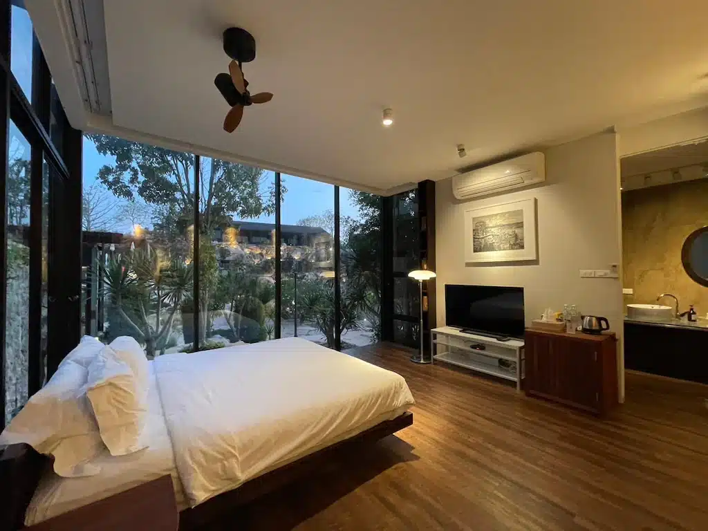 ห้องนอนสมัยใหม่พร้อมหน้าต่างบานใหญ่จากพื้นจรดเพดาน เตียงคู่ พื้นไม้ โทรทัศน์ เครื่องปรับอากาศ และวิวสวน—เหมาะสำหรับการพักผ่อนหลังจากสำรวจสถานที่ท่องเที่ยวเขาใหญ่.