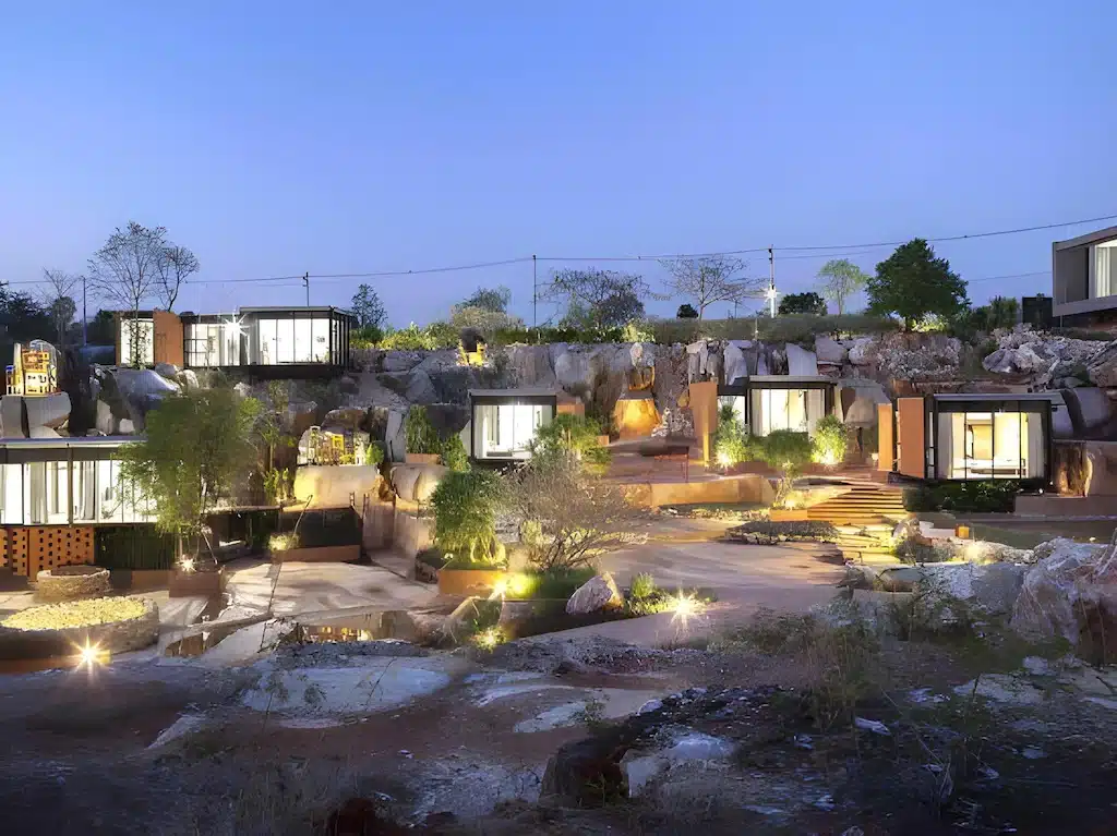 บ้านสไตล์โมเดิร์นและมินิมอลที่มีหน้าต่างขนาดใหญ่ถูกสร้างขึ้นในทิวทัศน์หินใกล้กับสถานที่ท่องเที่ยวเขาใหญ่ ล้อมรอบด้วยต้นไม้และส่องสว่างด้วยไฟภายนอกในยามพลบค่ำ.