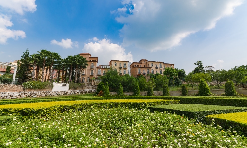 สวนที่ได้รับการตกแต่งอย่างประณีตด้วยพุ่มไม้รูปทรงเรขาคณิตและไม้ดอกหลากสีสัน ประดับประดาอาคารขนาดใหญ่สีเบจภายใต้ท้องฟ้าที่มีเมฆบางส่วน—เหมาะอย่างยิ่งสำหรับผู้ที่มองหา เขาใหญ่ที่เที่ยว หรือ สถานที่ท่องเที่ยวเขาใหญ่ที่น่าจดจำ.