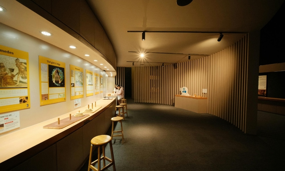 พื้นที่จัดแสดงในพิพิธภัณฑ์ที่มีแสงสว่างเพียงพอ พร้อมเก้าอี้สตูล, หน้าจอแสดงข้อมูลแบบโต้ตอบบนเคาน์เตอร์, โปสเตอร์ข้อมูลบนผนัง, และบุคคลที่นั่งอยู่เบื้องหลัง—เหมาะสำหรับผู้ที่ต้องการสถานที่ท่องเที่ยวเขาใหญ่ที่มีแหล่งท่องเที่ยวเชิงการศึกษา.