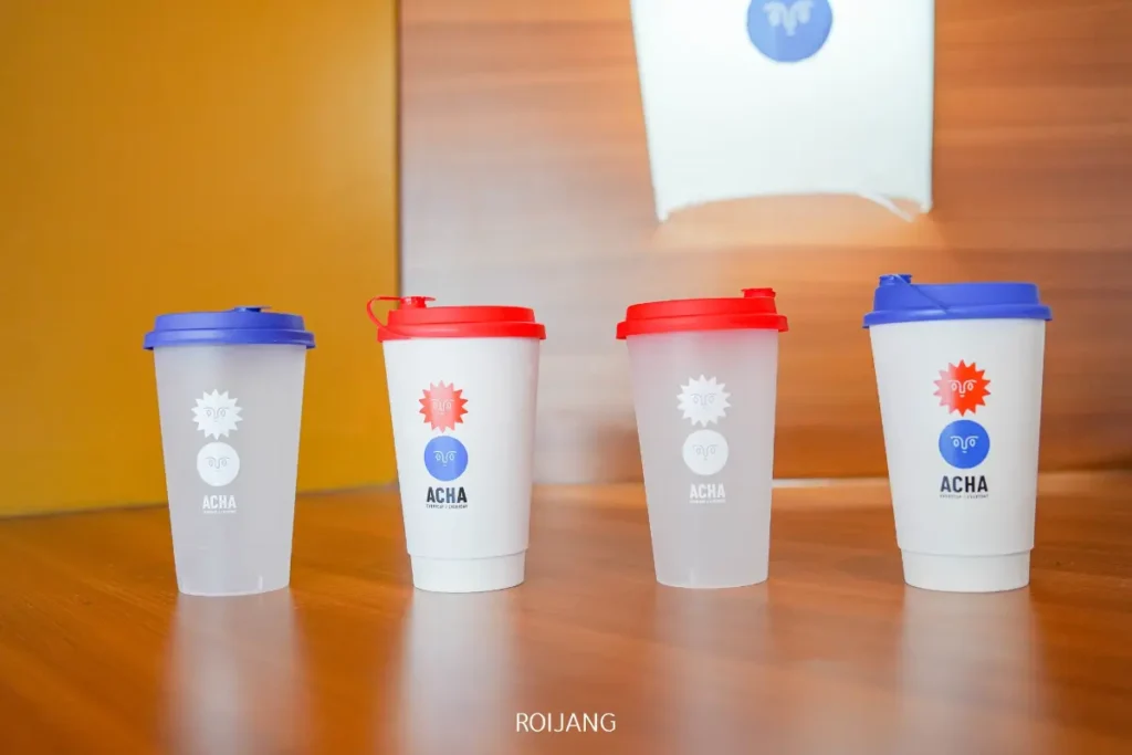 แก้วพลาสติกพร้อมฝาปิดสี่ใบ—สองใบสีน้ำเงิน สองใบสีแดง—ตั้งอยู่บนพื้นผิวไม้ แต่ละใบมีโลโก้และดีไซน์ "ACHA ภูเก็ต" พิมพ์อยู่ด้านหน้า.