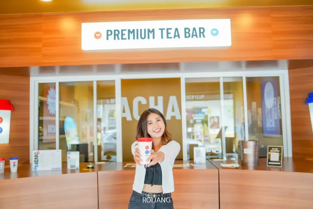 ผู้หญิงคนหนึ่งยืนอยู่ที่เคาน์เตอร์หน้าป้าย "Premium Tea Bar" ที่ ACHA ภูเก็ต ยิ้มและยื่นแก้วสำหรับซื้อกลับบ้านไปทางกล้อง.