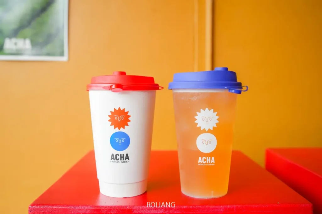 แก้วเครื่องดื่มสองใบที่มีแบรนด์ ACHA หนึ่งใบมีฝาสีแดงสำหรับเครื่องดื่มร้อนและอีกใบมีฝาสีฟ้าสำหรับเครื่องดื่มเย็น วางอยู่บนโต๊ะสีแดงที่ติดกับผนังสีส้มที่ ACHA ภูเก็ต.