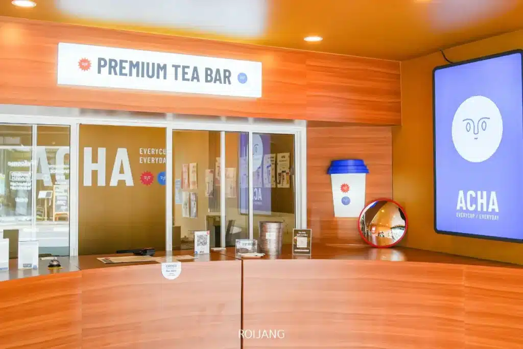 ภายในร้านชาที่มีป้าย "Premium Tea Bar" เคาน์เตอร์ไม้ตกแต่งด้วยของแบรนด์ และหน้าจอขนาดใหญ่แสดงโลโก้ "ACHA EVERYDAY EVERYDAY" สะท้อนบรรยากาศที่เชิญชวนของ ACHA ภูเก็ต.