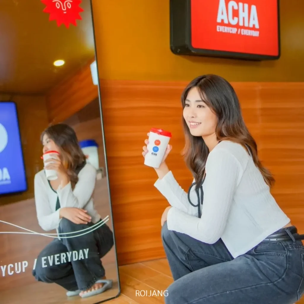 ผู้หญิงคนหนึ่งนั่งยองอยู่ภายในบ้าน ถือแก้วเครื่องดื่มจากร้าน ACHA ภูเก็ต และยิ้มให้กับภาพสะท้อนของตัวเองในกระจกบานใหญ่.