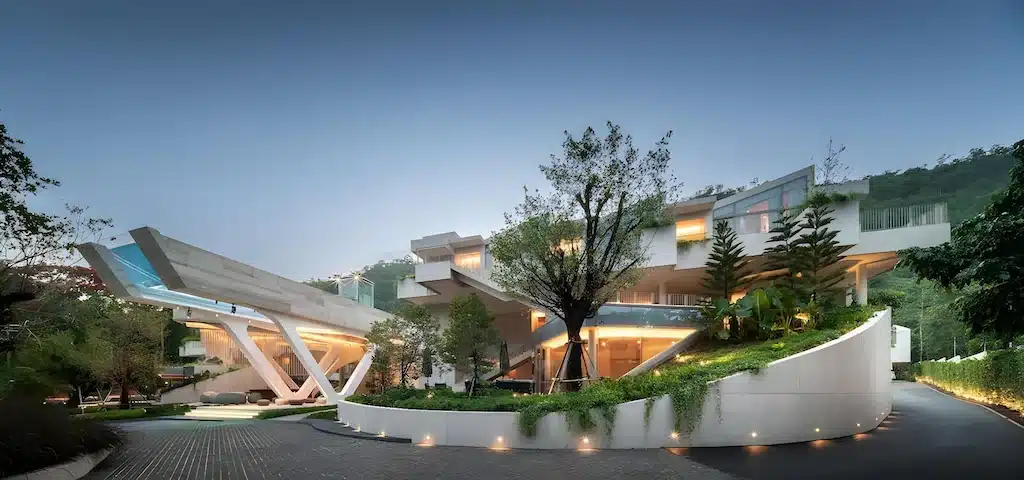 บ้านหลายชั้นสมัยใหม่พร้อมหน้าต่างกระจกขนาดใหญ่ การออกแบบเรขาคณิต ไฟภายนอก สวนภูมิทัศน์ และโครงสร้างยื่นที่น่าทึ่ง—สถานที่พักผ่อนในอุดมคติท่ามกลางต้นไม้และเนินเขาในยามพลบค่ำใกล้แหล่งท่องเที่ยวเขาใหญ่.