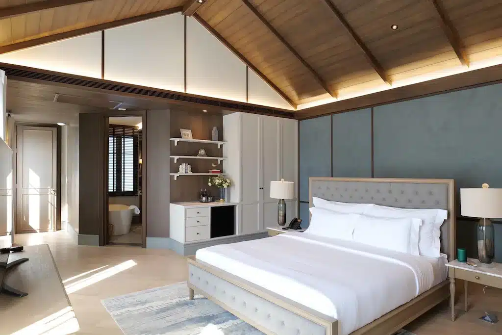 ห้องนอนที่กว้างขวางและทันสมัยพร้อมเตียงขนาดใหญ่, เพดานไม้, โคมไฟข้างเตียง, ชั้นวางของแบบบิวท์อิน, และประตูเปิดโล่งไปยังห้องน้ำ—เหมาะสำหรับการผ่อนคลายหลังจากวันที่ออกไปเที่ยวเขาใหญ่.