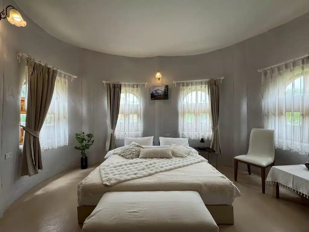 เตียงที่จัดไว้อย่างเรียบร้อยในห้องทรงกลมที่มีหน้าต่างโค้งสี่บาน ผ้าม่านบางเบา เก้าอี้หนึ่งตัว และการตกแต่งโทนสีเรียบ—เหมาะอย่างยิ่งสำหรับการพักผ่อนหลังจากเที่ยวชมแหล่งท่องเที่ยวเขาใหญ่.