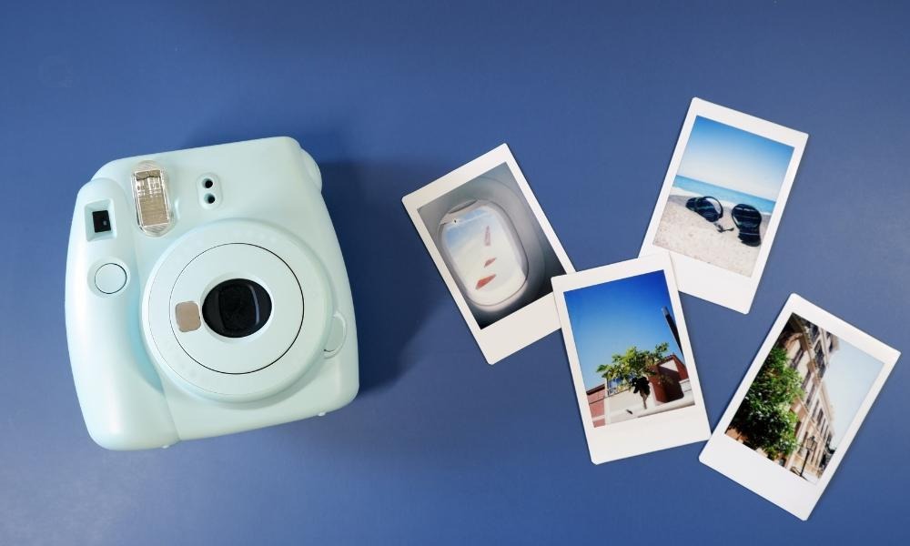 กล้องถ่ายรูปอินสแตนท์สีฟ้าอ่อนวางอยู่บนพื้นผิวสีฟ้าข้างๆ รูปถ่ายอินสแตนท์สี่ใบ แต่ละใบแสดงฉากที่แตกต่างกัน.