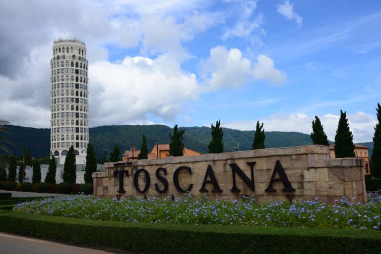 ป้ายหินที่เขียนว่า "Toscana" ตั้งอยู่หน้าดอกไม้ที่จัดแต่งอย่างสวยงาม พร้อมด้วยหอเอนเมืองปิซาจำลองและเนินเขาเป็นฉากหลัง—เหมาะสำหรับผู้ที่มองหาสถานที่ท่องเที่ยวเขาใหญ่ภายใต้ท้องฟ้าที่มีเมฆบางส่วน.