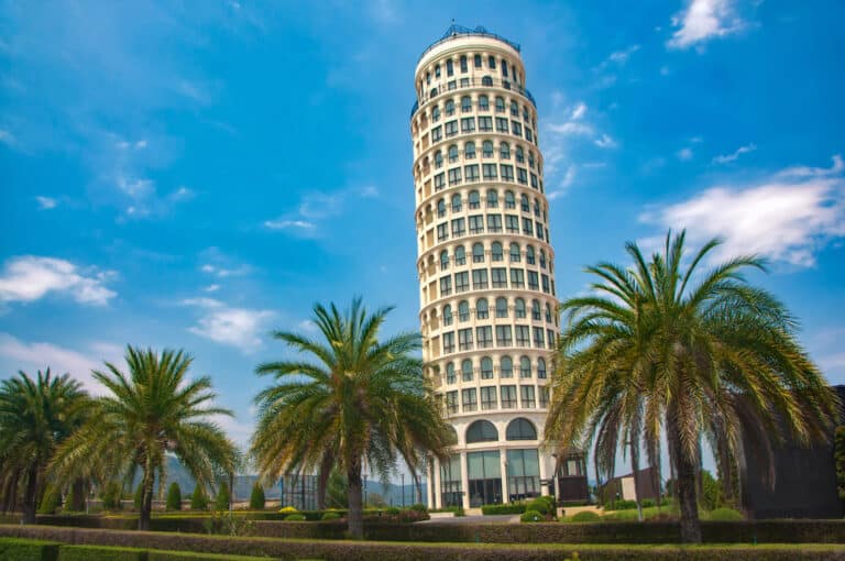 อาคารทรงกระบอกสูงที่มีหน้าต่างโค้งหลายบานตั้งอยู่ท่ามกลางต้นปาล์มใต้ท้องฟ้าสีครามที่มีเมฆกระจายอยู่ จับภาพเสน่ห์อันเป็นเอกลักษณ์ของ สถานที่ท่องเที่ยวเขาใหญ่.