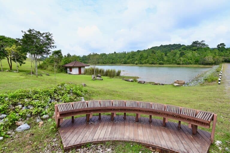 ม้านั่งไม้โค้งทอดยาวมองเห็นทะเลสาบเล็กๆ ที่มีหญ้า ต้นไม้ อาคารสีขาว และท้องฟ้าที่มีเมฆครึ้ม—เหมาะสำหรับการพักผ่อนที่ ที่เที่ยวเขาใหญ่ หนึ่งใน สถานที่ท่องเที่ยวเขาใหญ่ ที่มีเสน่ห์.