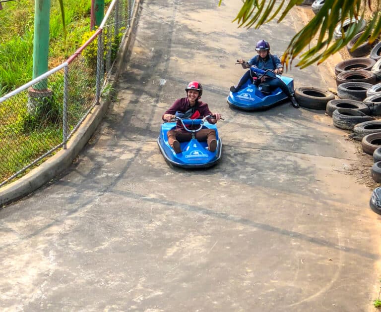 สองคนสวมหมวกกันน็อคขี่โกคาร์ทไฟฟ้าสีน้ำเงินบนสนามปูพื้นกลางแจ้งใกล้กองยางและรั้วต้นไม้—กิจกรรมสนุกสำหรับผู้ที่มองหาตัวเลือกท่องเที่ยวเขาใหญ่.