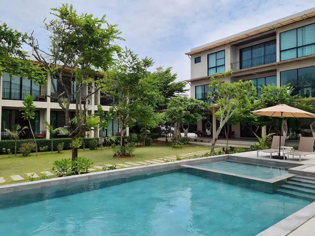 โรงแรมสมัยใหม่ โรงแรมขนอม มีหน้าต่างบานใหญ่ที่มองเห็นสระว่ายน้ำรูปสี่เหลี่ยมผืนผ้าและสวนที่จัดแต่งอย่างสวยงามพร้อมต้นไม้และเก้าอี้พักผ่อนใต้ร่ม.