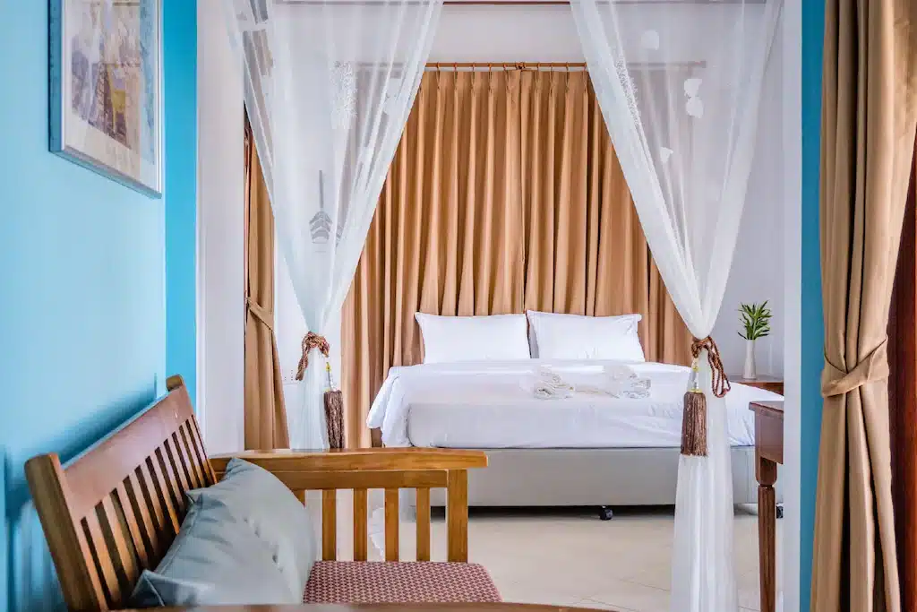 เตียงที่ปูเรียบร้อยด้วยผ้าปูที่นอนสีขาวสะอาดตาถูกโอบล้อมด้วยผ้าม่านบางเบา ม้านั่งไม้พร้อมเบาะรองนั่งตั้งอยู่ด้านหน้า และผ้าม่านสีเบจคลุมหน้าต่าง สร้างบรรยากาศที่เงียบสงบเหมาะสำหรับการเข้าพักของคุณที่ รีสอร์ทขนอม.