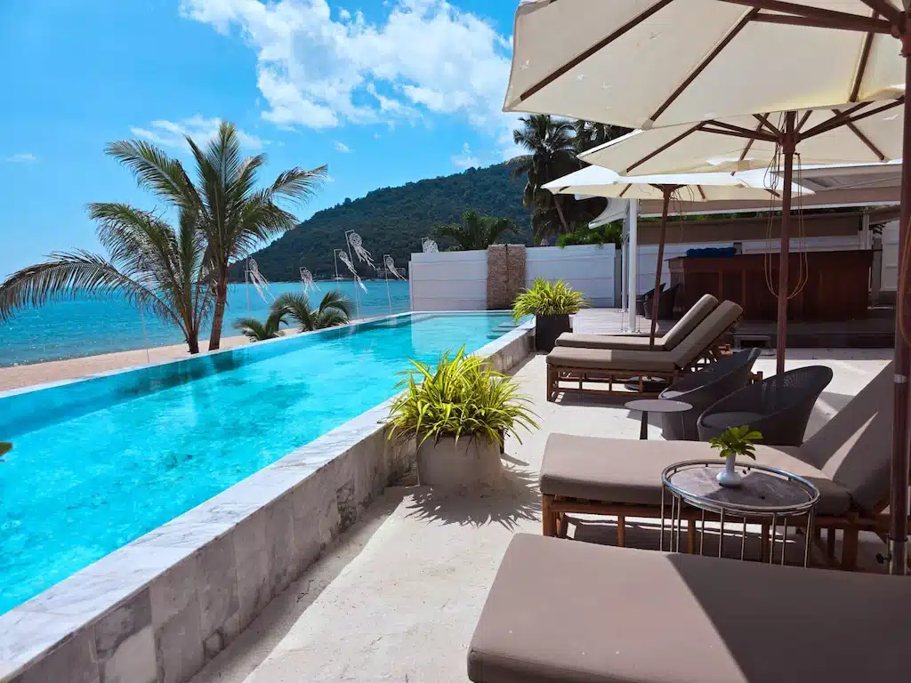 พื้นที่เลานจ์ริมสระว่ายน้ำกลางแจ้งพร้อมเตียงอาบแดด ร่ม และบาร์ ณ รีสอร์ทในขนอมที่มองเห็นชายหาดและมหาสมุทร โดยมีต้นปาล์มและเนินเขาเป็นฉากหลังภายใต้ท้องฟ้าสีคราม.