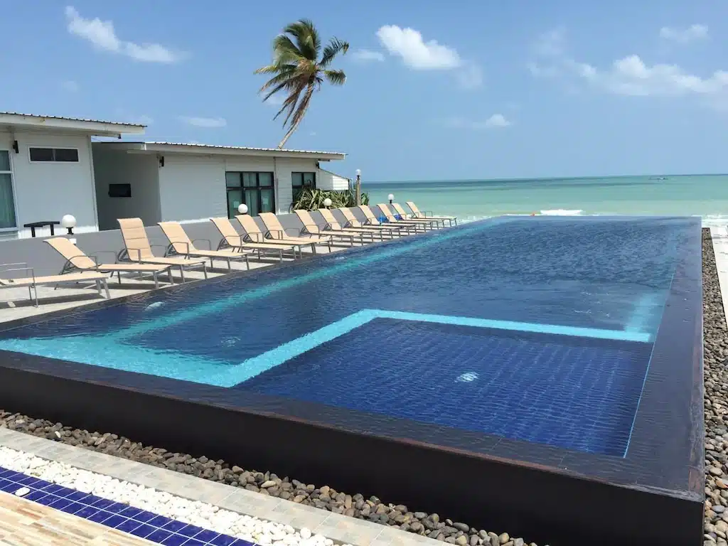 สระว่ายน้ำอินฟินิตี้ทรงสี่เหลี่ยมผืนผ้าพร้อมกระเบื้องสีน้ำเงิน ล้อมรอบด้วยเก้าอี้อาบแดดข้างอาคารสมัยใหม่ ณ ที่พักที่สวยงามในขนอม มองเห็นชายหาดและมหาสมุทรภายใต้ท้องฟ้าที่มีเมฆบางส่วน.