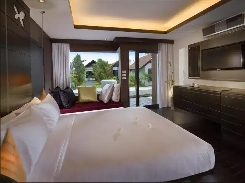 ห้องนอนสมัยใหม่พร้อมเตียงขนาดใหญ่, ที่นั่งริมหน้าต่างที่แสนสบาย, โทรทัศน์จอแบน, และวิวของสวนและวิลล่าผ่านประตูกระจก—เหมาะสำหรับการผ่อนคลายที่ ที่พักขนอม.